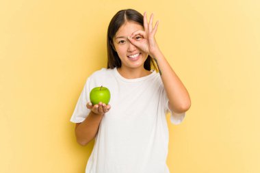 Sarı arka planda yalıtılmış elma yiyen genç Asyalı kadın heyecanlı..