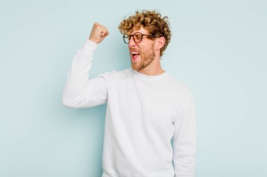 Mavi arka planda izole edilmiş genç beyaz bir adam zaferi, tutkuyu ve coşkuyu kutluyor, mutlu bir ifade..