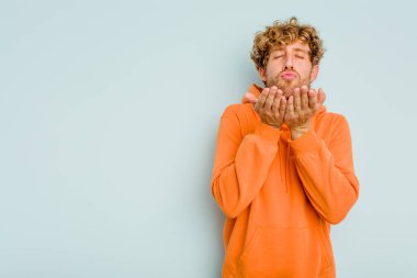 Kafkasyalı genç adam mavi arka planda izole edilmiş dudaklar kıvrılıyor ve hava öpücüğü göndermek için avuç içleri tutuyor..