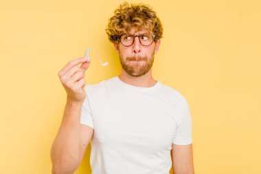 Sarı arka planda izole edilmiş işitme cihazı takan genç beyaz adam şaşkın ve güvensiz hissediyor..