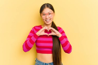 Sarı arka planda izole edilmiş genç Asyalı kadın gülümsüyor ve elleriyle kalp şeklini gösteriyor..