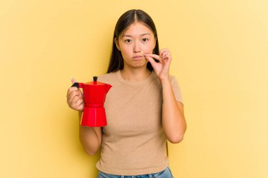 Sarı arka planda kahve makinesini tutan genç Asyalı kadın dudaklarında parmaklarıyla bir sır saklıyor..
