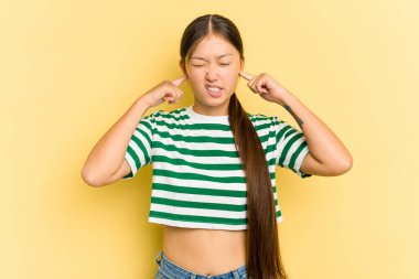 Sarı arka planda izole edilmiş genç Asyalı kadın kulaklarını parmaklarıyla kapatıyor, gürültülü bir ortam yüzünden stresli ve çaresiz..