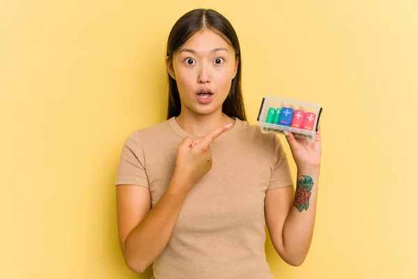 電池を持っている若いアジアの女性は 側面を指す黄色の背景に隔離されたそれらをリサイクルする — ストック写真