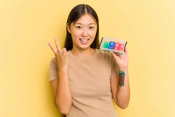 黄色の背景にバッテリーを保持している若いアジアの女性は喜びと屈託のない指で平和のシンボルを示す上で隔離されたリサイクル — ストック写真