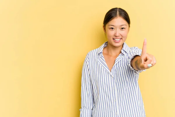 在黄色背景下被隔离的年轻亚洲女人用手指表现出第一位 — 图库照片