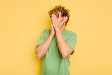 Sarı arka planda izole edilmiş genç beyaz adam parmaklarının arasından korkmuş ve gergin bir şekilde göz kırp..
