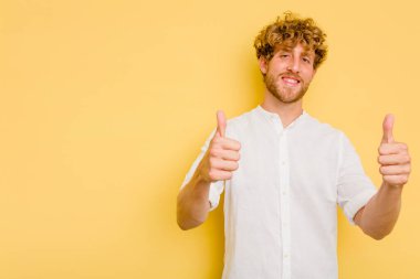 Sarı arka planda izole edilmiş genç beyaz adam gülümsüyor ve başparmağını kaldırıyor.