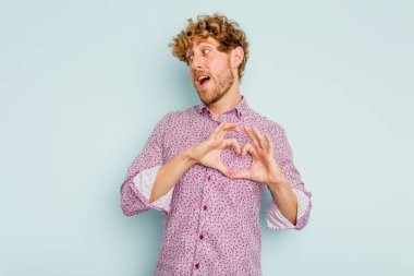 Mavi arka planda izole edilmiş genç beyaz adam gülümsüyor ve elleriyle kalp şeklini gösteriyor..