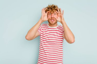 Mavi arka planda izole edilmiş genç beyaz adam bir başarı fırsatı bulmak için gözlerini açık tutuyor..