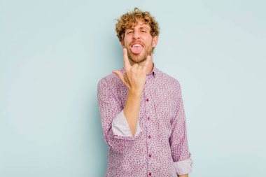 Mavi arka planda izole edilmiş genç beyaz adam parmaklarıyla kaya hareketi gösteriyor.