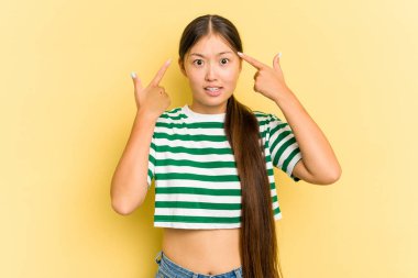 Sarı arka planda izole edilmiş genç bir Asyalı kadın işaret parmağıyla hayal kırıklığı gösteriyor..