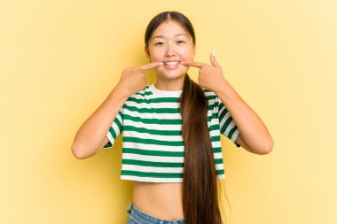 Sarı arka planda izole edilmiş genç Asyalı kadın gülümsüyor, parmaklarını ağıza işaret ediyor..