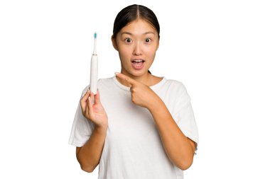 Elinde elektrikli diş fırçası olan genç bir Asyalı kadın yeşil renkli arka planda izole edilmiş. Yan tarafı işaret ediyor.