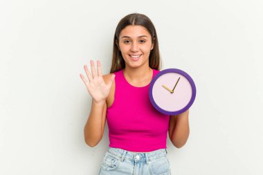 Genç Hintli bir kadın beyaz arka planda izole edilmiş bir saat tutuyor. Beş numarayı parmaklarıyla gülümsüyor..