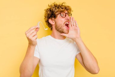 Sarı arka planda izole edilmiş işitme cihazı takan genç beyaz adam bağırıyor ve avuç içini açık tutuyor..