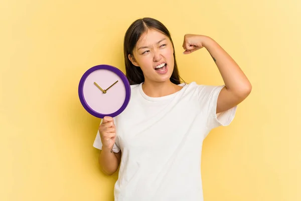 Joven Asiática Sosteniendo Reloj Aislado Sobre Fondo Amarillo Levantando Puño —  Fotos de Stock