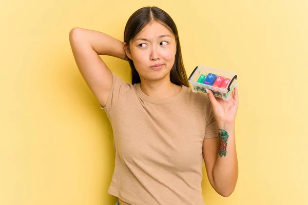Young Asian woman holding batteries to recycle them isolated on yellow background touching back of head, thinking and making a choice.