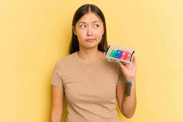 黄色の背景の混乱に隔離されたそれらをリサイクルするために電池を保持している若いアジアの女性は 疑問と不確定な感じ — ストック写真