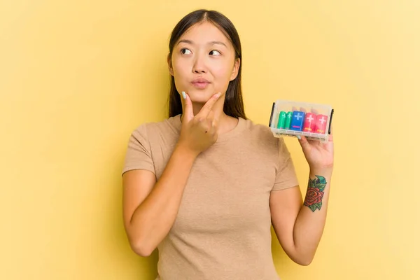 電池を持っている若いアジアの女性は 疑わしく懐疑的な表情で横に見える黄色の背景に隔離されたそれらをリサイクルする — ストック写真