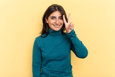 Sarı arka planda izole edilmiş genç Hintli kadın iki numarayı parmaklarıyla gösteriyor..