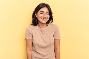 Sarı arka planda izole edilmiş genç Hintli kadın gülüyor ve gözlerini kapatıyor, rahatlamış ve mutlu hissediyor..