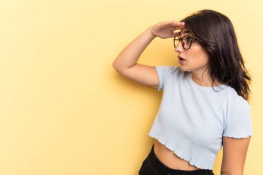 Sarı arka planda izole edilmiş genç Hintli kadın uzaklara bakıyor ve elini alnına koyuyor..