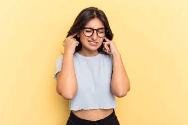 Sarı arka planda izole edilmiş genç bir Hintli kadın parmaklarıyla kulaklarını kapatıyor, gürültülü bir ortam yüzünden stresli ve çaresiz..
