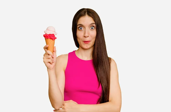 年轻的高加索女人吃着冰淇淋 孤立地耸了耸肩 睁开眼睛迷惑不解 — 图库照片