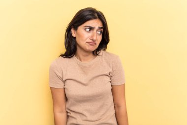 Sarı arka planda izole edilmiş genç Hintli kadın omuzlarını silker ve gözleri açık kalır..