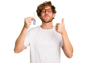 Beyaz arka planda izole edilmiş, gülümseyen ve baş parmağını kaldıran ev anahtarlarını elinde tutan genç beyaz bir adam.