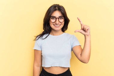 Sarı arka planda izole edilmiş genç bir Hintli kadın işaret parmaklarıyla küçük bir şey tutuyor, gülümsüyor ve kendinden emin..