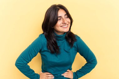 Sarı arka planda izole edilmiş genç Hintli kadın ellerini kalçasında tutuyor..