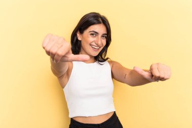 Sarı arka planda izole edilmiş genç Hintli kadın iki parmağını da kaldırıyor, gülümsüyor ve kendinden emin..