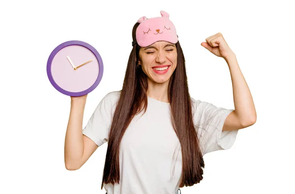 Young Caucasian Woman Wearing Pajama Holding Clock Raising Fist Victory — Stock Photo, Image