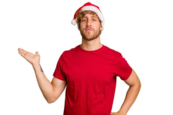 白い背景に隔離されたクリスマスのサンタの帽子をかぶっている若い白人男性は 手のひらにコピースペースを示し 腰に別の手を握っています — ストック写真