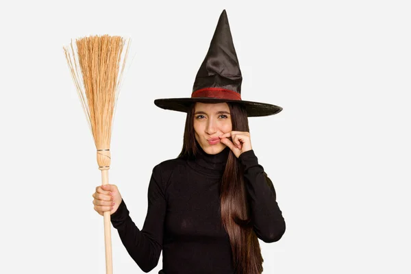 年轻的高加索女人穿着巫婆的衣服 手里拿着一把用手指捂住嘴唇的扫帚保守秘密 — 图库照片