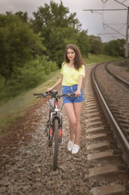 Bir kız ve bisikleti. Sarı tişörtlü ve kot şortlu yakışıklı bir genç bisikletle şehrin dışında yürüyor. Bir fotoğrafçıya poz veriyor..