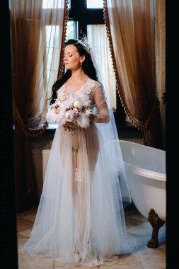 Gelin, şeffaf bir elbise ve iç çamaşırı içinde, elinde bir tüyle eski bir banyonun yanında oturuyor, gelinin sabahında..