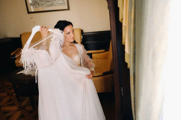 a bride dressed in a boudoir transparent dress and underwear holds her wedding dress in her hands in the interior of the house.