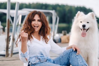 Büyük beyaz bir köpeği olan mutlu bir kadın gün batımında denizin yakınındaki bir rıhtımda yatıyor..