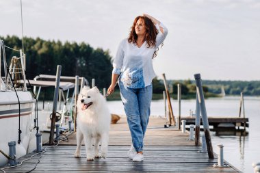 Büyük beyaz köpekli mutlu bir kadın yatın ve denizin yakınındaki iskelede yürüyor..