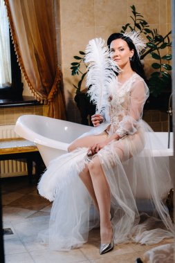 Gelin, şeffaf bir elbise ve iç çamaşırı giyip, eski bir küvete oturmuş, elinde bir tüyle, gelinin sabahında..