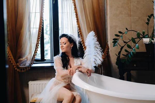 Gelin, şeffaf bir elbise ve iç çamaşırı içinde, elinde bir tüyle eski bir banyonun yanında oturuyor, gelinin sabahında..