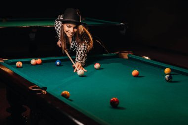 Bilardo kulübünde elinde ıstakayla şapkalı bir kız topa vuruyor. Bilardo oynuyor..