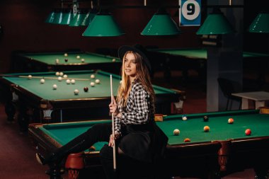 Bilardo kulübünde şapkalı bir kız elinde ıstakayla bilardo masasında oturuyor. Bilardo oynuyor..