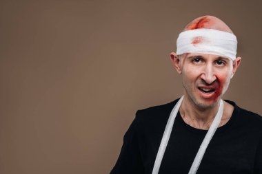 Kafası bandajlı ve kolunda alçı olan hırpalanmış bir adam gri bir arka planda duruyor..