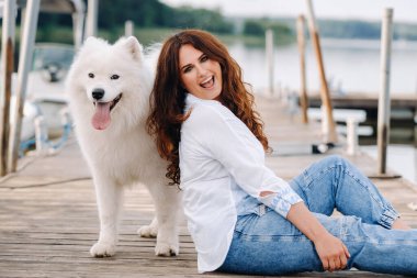 Büyük beyaz bir köpeği olan mutlu bir kadın gün batımında deniz kenarında bir rıhtımda oturur..