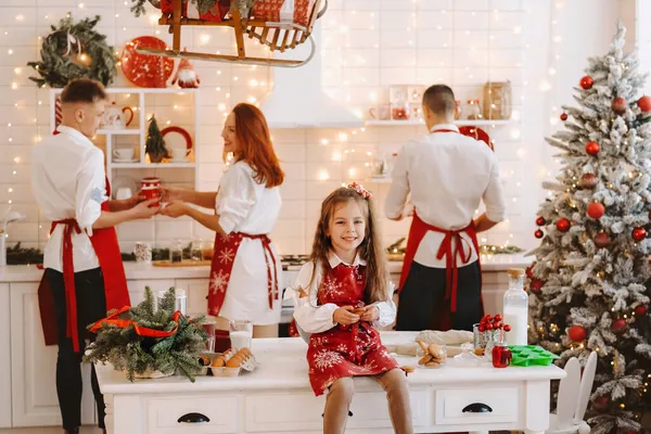 新年厨房里 一个小女孩手里拿着饼干坐在桌子上 她的父母正在准备圣诞大餐 — 图库照片