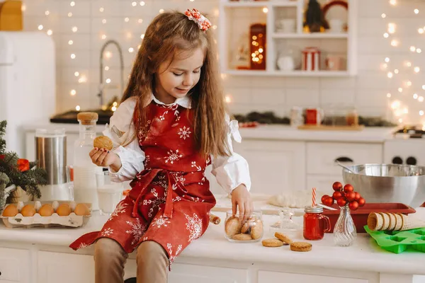 圣诞节厨房里一个快乐的小女孩手里拿着饼干坐在桌子上 — 图库照片